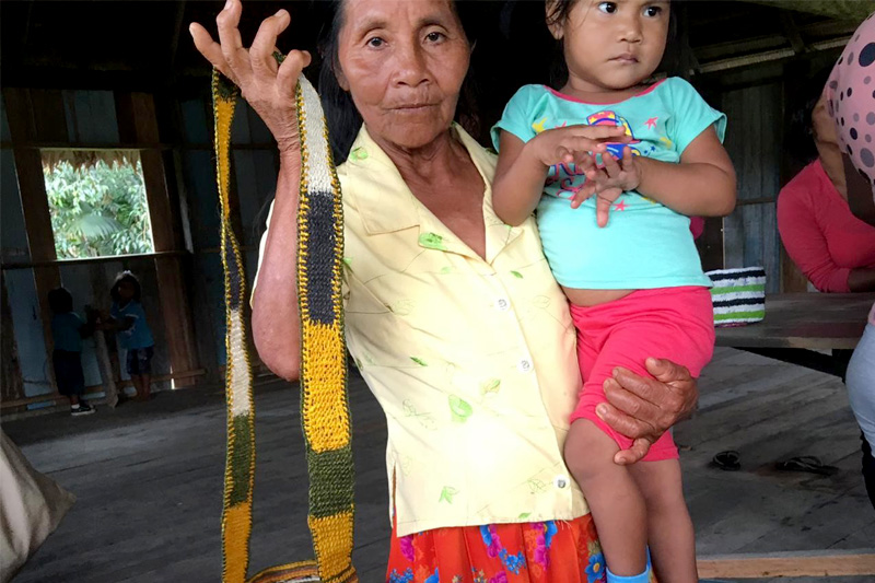 Mujer murui sostiene a una niña murui y muestra una mochila, para resaltar la importancia de enseñar sobre tejidos y colores.